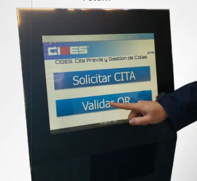 Mano pulsando «Validar QR» en la pantalla táctil de un tótem CIGES